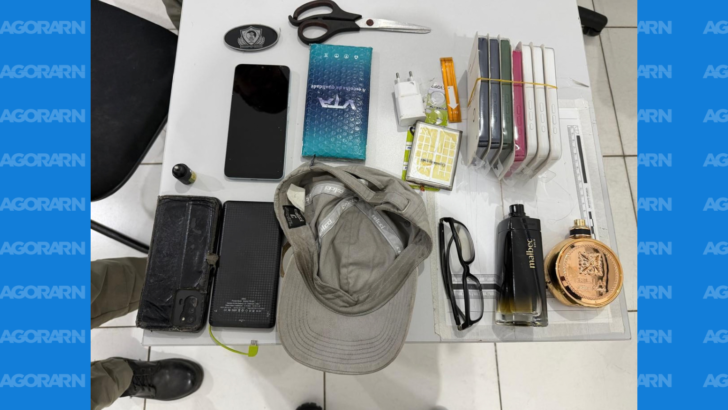Celulares, boné, óculos, perfumes, tesoura e outros objetos apreendidos estão dispostos sobre uma mesa