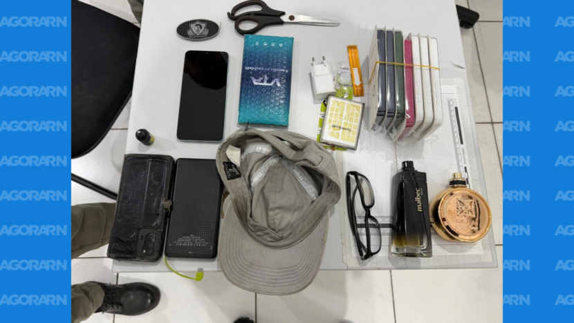 Celulares, boné, óculos, perfumes, tesoura e outros objetos apreendidos estão dispostos sobre uma mesa