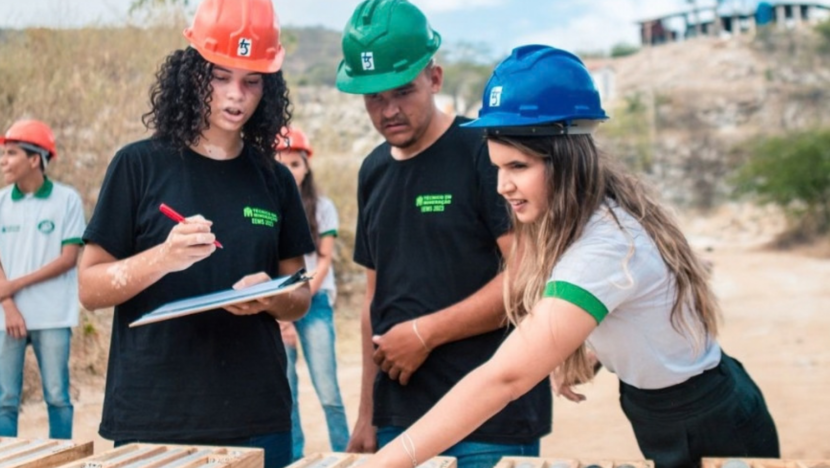Três jovens usando capacetes observam e fazem anotações sobre amostras dispostas em caixas durante atividade de campo