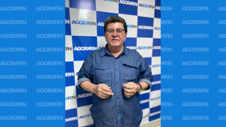 Homem de óculos e camisa azul posa diante de painel com a marca Agora RN