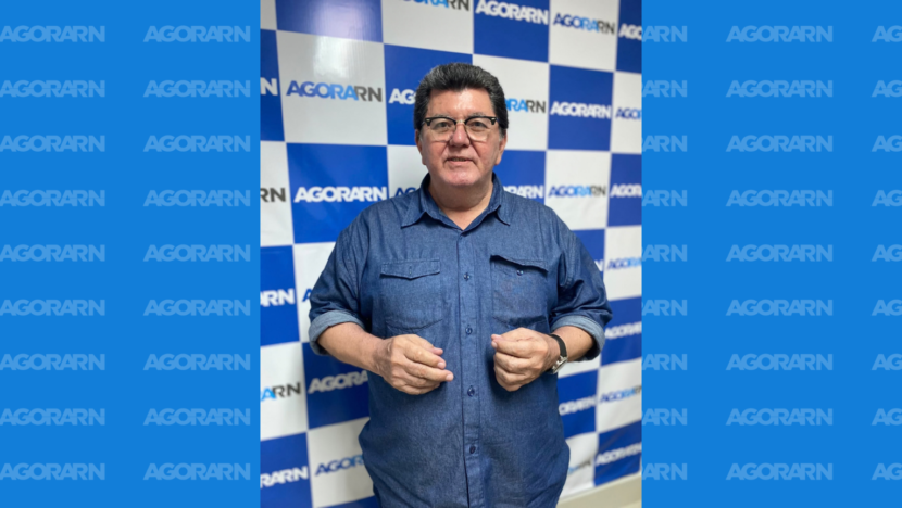 Homem de óculos e camisa azul posa diante de painel com a marca Agora RN