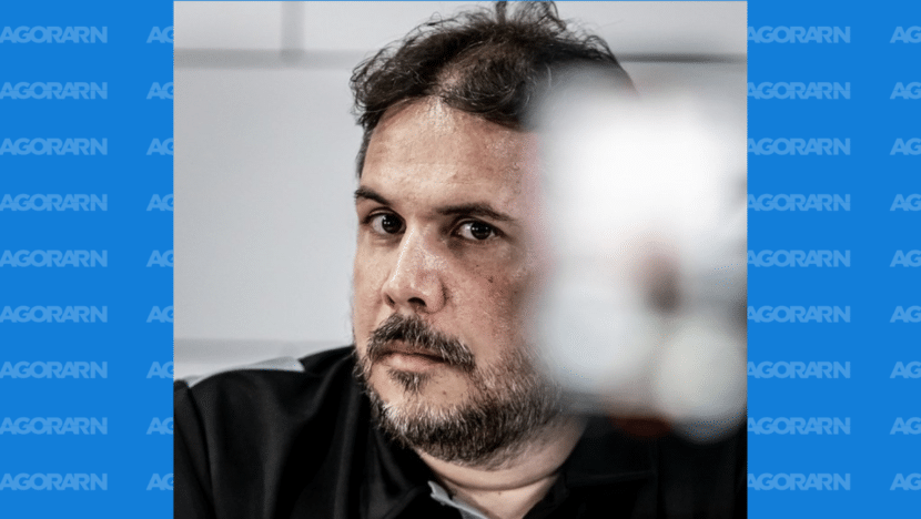 Gustavo Cartaxo em close, com barba e cabelo curto, olhando para o lado, com parte da imagem desfocada em primeiro plano.