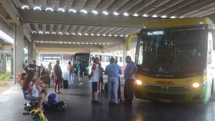 Passageiros com bagagens esperam na plataforma coberta da rodoviária de Natal, ao lado de um ônibus intermunicipal amarelo e verde com o letreiro Via Macau