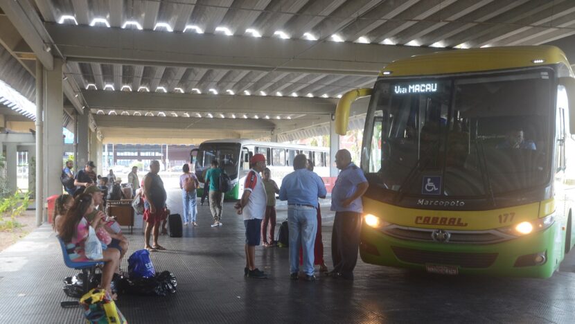Passageiros com bagagens esperam na plataforma coberta da rodoviária de Natal, ao lado de um ônibus intermunicipal amarelo e verde com o letreiro Via Macau
