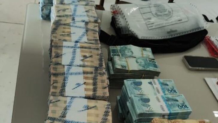 Maços de notas de R$ 100 e R$ 50, alguns lacrados com etiqueta de banco, empilhados sobre uma mesa ao lado de uma bolsa escura e de um envelope de evidência