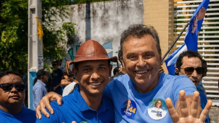 Carlos Eduardo Alves e Allyson Bezerra, de camisa azul, abraçados e sorrindo durante ato de campanha na rua, com adesivos dos números 44 e 555