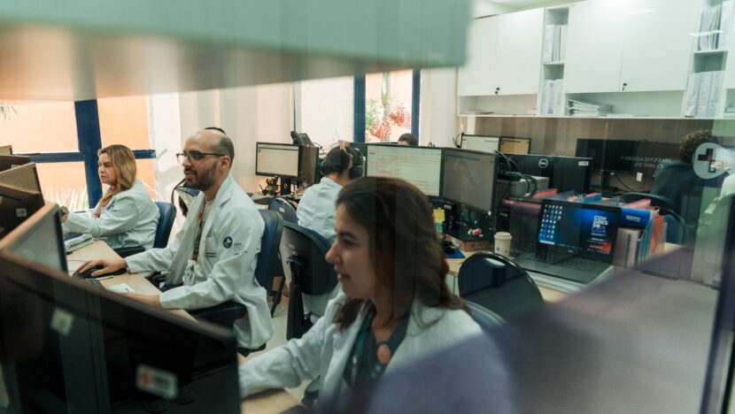 Profissionais de jaleco branco trabalham em computadores na sala do Centro de Pesquisa Clínica da Liga contra o Câncer, vistos através de um vidro com a inscrição Investigadores Clínicos