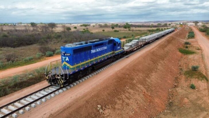Locomotiva azul da Transnordestina puxa vagões carregados de dormentes de concreto sobre um trecho de via em construção, em meio à caatinga, com estrada de terra ao lado