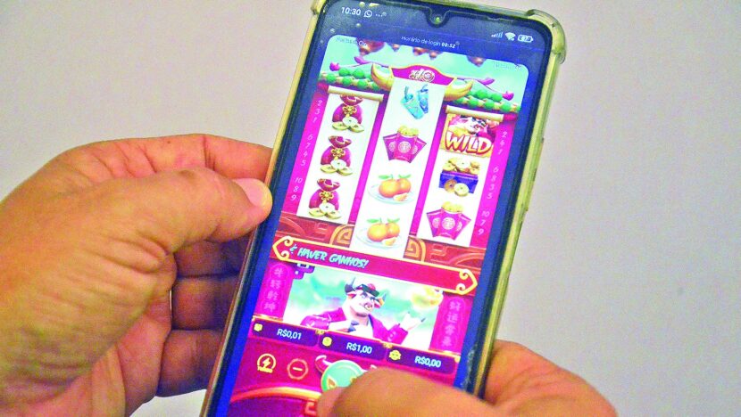 Mãos seguram um celular com a tela de um jogo de cassino online do tipo caça-níquel, com rolos de símbolos, o botão Wild e valores de aposta em reais