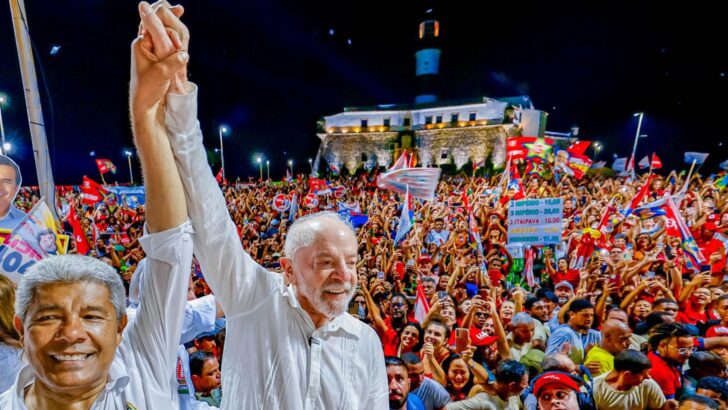 Lula e Jerônimo Rodrigues, de mãos dadas e braços erguidos, diante de uma multidão com bandeiras vermelhas em ato noturno de campanha junto ao Farol da Barra, em Salvador
