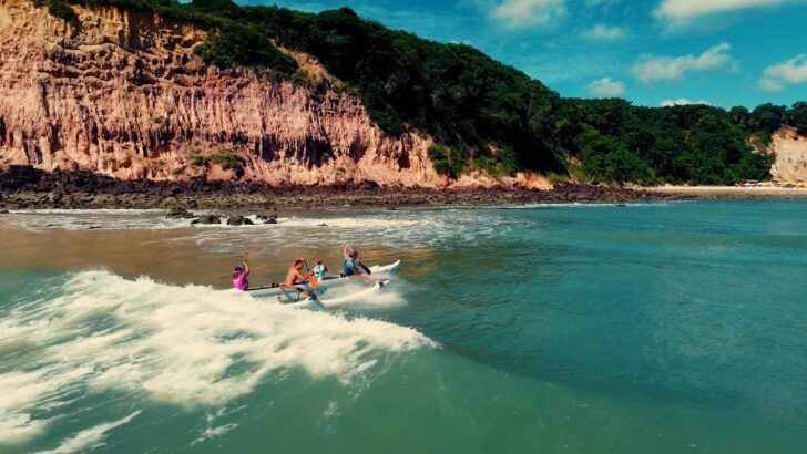 Quatro remadores conduzem uma canoa havaiana sobre a espuma de uma onda junto à costa, diante das falésias avermelhadas e da mata de Pipa