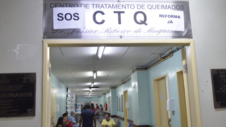 Entrada do Centro de Tratamento de Queimados do Hospital Walfredo Gurgel, com cartazes de SOS sobre a placa e pacientes no corredor