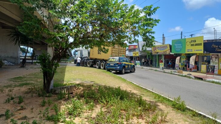 Carreta parada em via urbana com veículo da Polícia Rodoviária Federal próximo e estabelecimentos comerciais ao fundo