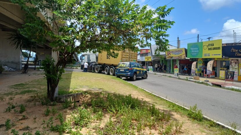 Carreta parada em via urbana com veículo da Polícia Rodoviária Federal próximo e estabelecimentos comerciais ao fundo