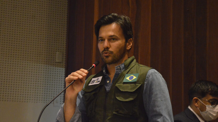 Fábio Faria fala ao microfone diante de painel de madeira, de colete verde sobre camisa jeans, com a bandeira do Brasil no bolso