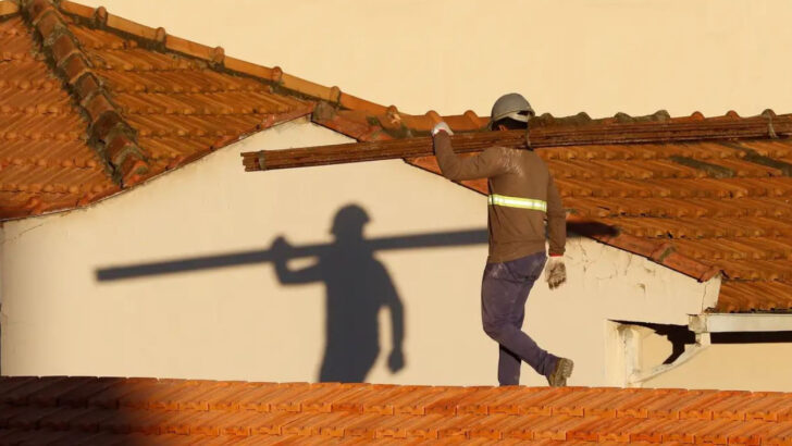 Trabalhador da construção civil carrega viga sobre telhado residencial