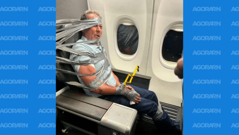 Passageiro sentado junto à janela de um avião, preso à poltrona com faixas de fita adesiva ao redor do tronco e da cabeça.