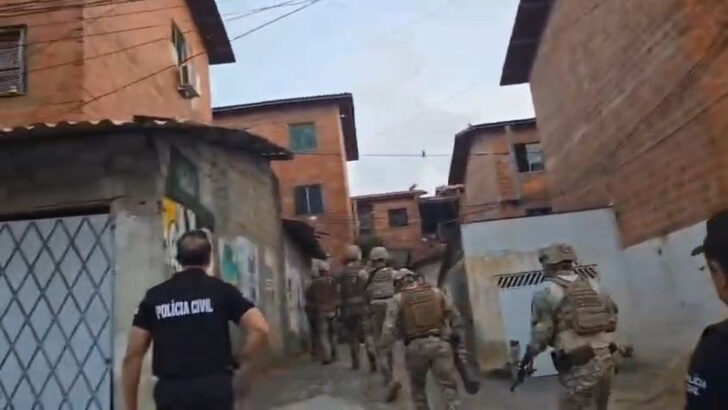 Agentes da Polícia Civil e equipe tática caminham por rua estreita durante cumprimento de mandados