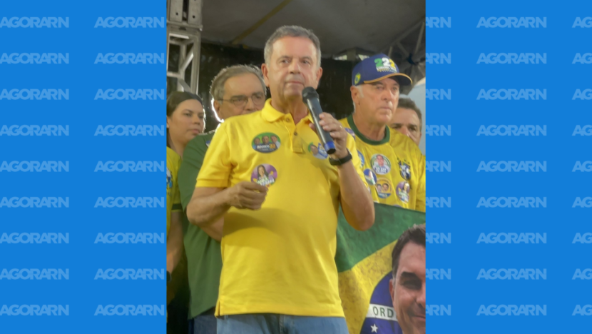 Rogério Marinho veste camisa amarela e segura um microfone durante manifestação, cercado por participantes com roupas nas cores do Brasil.