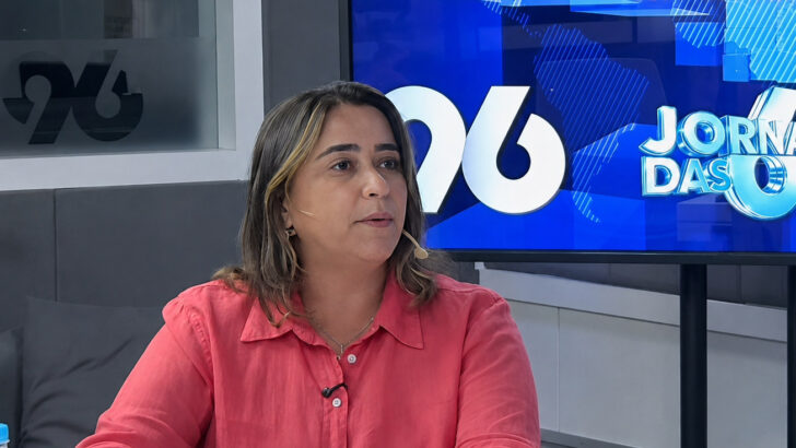 Samanda Alves durante entrevista em estúdio de rádio, com microfone de lapela e papéis sobre a mesa