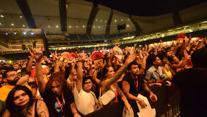 Público de mãos erguidas em frente ao palco durante show noturno em arena coberta