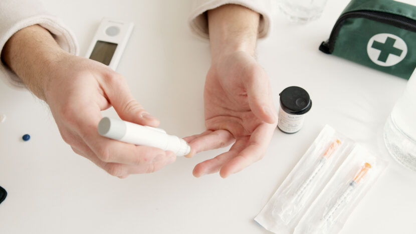 Pessoa faz teste de glicemia com lancetador ao lado de medidor, tiras de teste e seringas