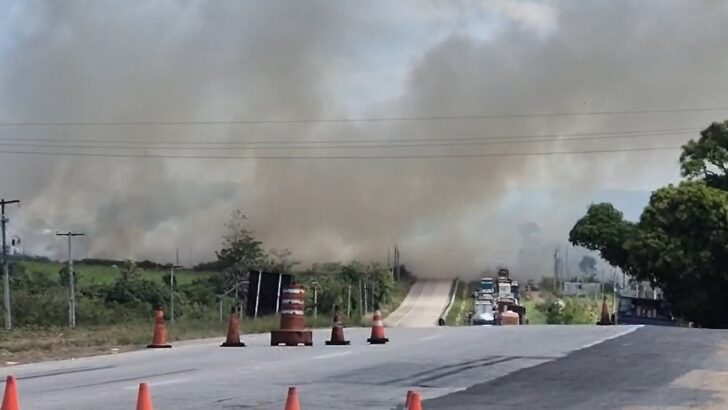 Trecho de rodovia parcialmente encoberto por fumaça, com cones na pista e veículos ao fundo