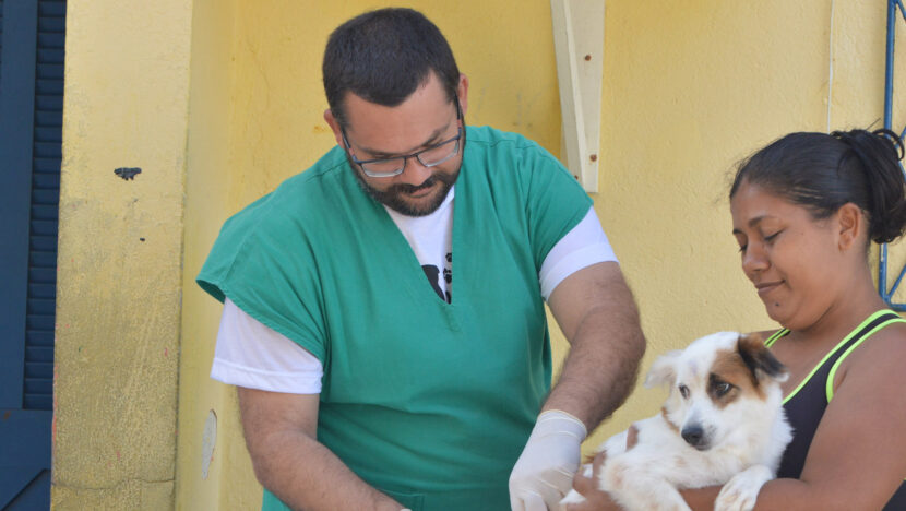 Profissional de jaleco verde e luvas prepara a vacina antirrábica enquanto uma mulher segura seu cão