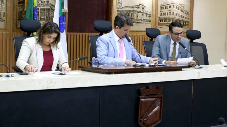 Uma vereadora e dois vereadores sentados à mesa com papéis e microfones no plenário da Câmara de Natal
