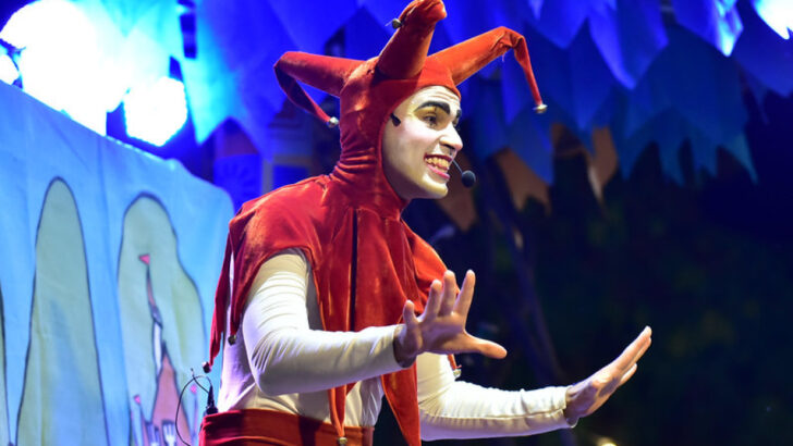 Artista usa figurino vermelho com chapéu de pontas e gesticula no palco