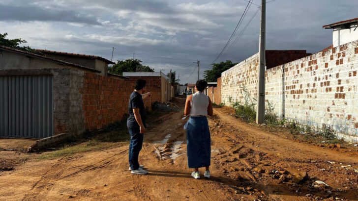 Duas pessoas em rua de terra, com trecho úmido e escavação junto à margem