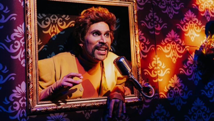Ator se apresenta dentro de uma moldura, diante de um microfone, em cena teatral