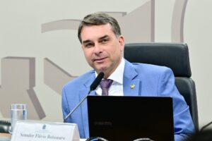 Homem de terno azul sentado à mesa, diante de microfone e computador, durante sessão
