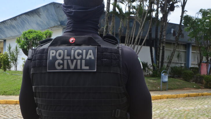 Policial civil visto de costas, com colete identificado, diante de prédio e árvores