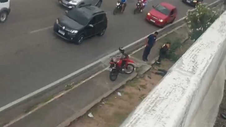 Imagem aérea de trecho da BR-101 com carros e motocicletas em circulação, uma motocicleta vermelha parada junto à calçada e pessoas próximas à via