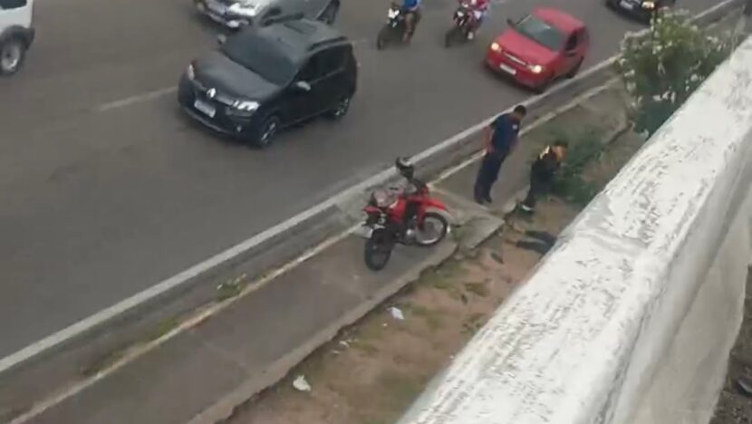 Imagem aérea de trecho da BR-101 com carros e motocicletas em circulação, uma motocicleta vermelha parada junto à calçada e pessoas próximas à via