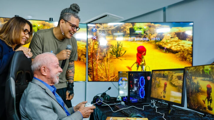 Lula sentado diante de monitores, segurando um controle de videogame, acompanhado por participantes do evento no Palácio do Planalto