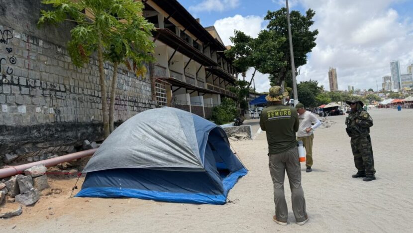 Barraca de camping azul e cinza instalada na areia da Praia de Ponta Negra, próxima a um muro, enquanto agentes de fiscalização ambiental e da Guarda Municipal acompanham a ocorrência