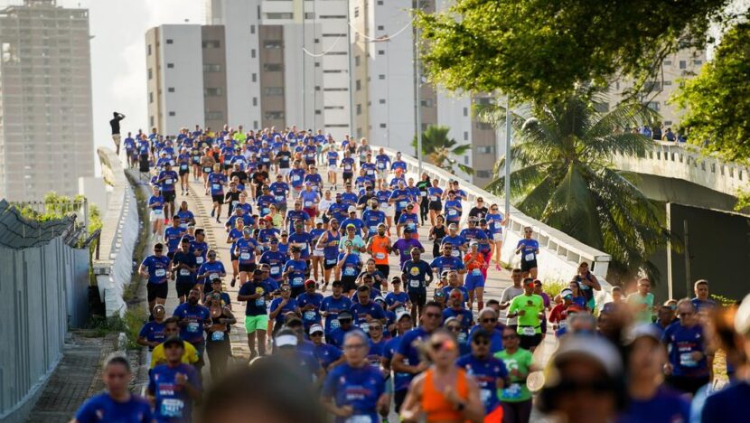 Grupo de corredores com números de peito percorre uma via em Natal, com prédios e árvores ao fundo