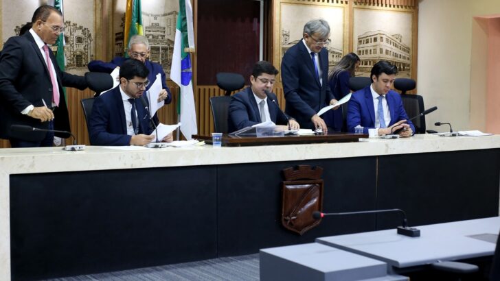 Vereadores sentados e em pé junto à mesa diretora da Câmara de Natal com documentos e microfones