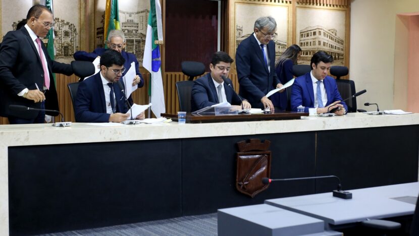 Vereadores sentados e em pé junto à mesa diretora da Câmara de Natal com documentos e microfones