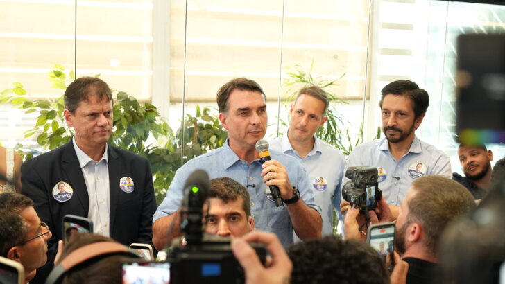Flávio Bolsonaro, de camisa azul-clara, fala ao microfone cercado por jornalistas e câmeras durante evento em São Paulo
