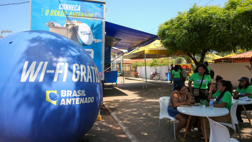 Mulheres com camisetas verdes atendem uma moradora em mesa ao ar livre, ao lado da unidade móvel e de um balão azul do Brasil Antenado