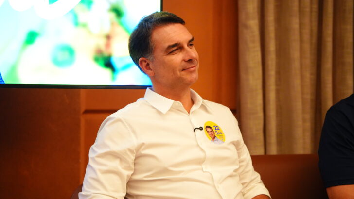 Flávio Bolsonaro sentado, usando camisa branca com adesivo de campanha, diante de cortinas