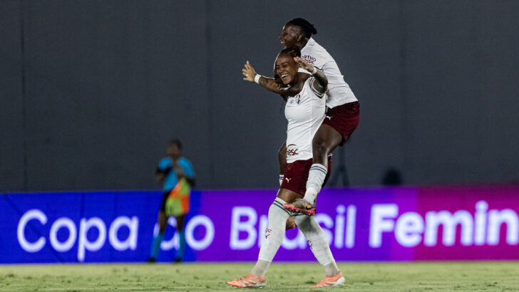 Duas jogadoras do Fluminense, com camisas brancas e shorts grenás, comemoram abraçadas no gramado do Luso Brasileiro