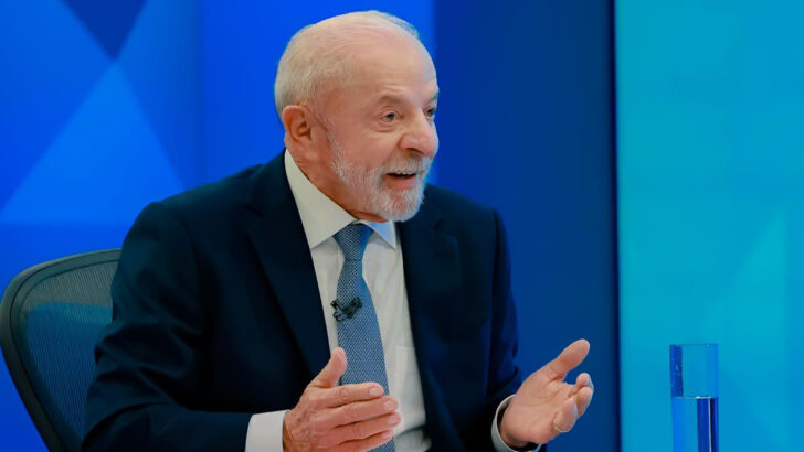 Lula, de terno azul e gravata, fala com as mãos abertas diante de uma mesa com papéis e copo de água, em estúdio de fundo azul