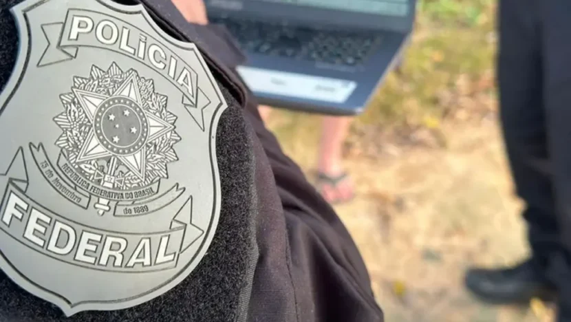 Imagem em primeiro plano de um distintivo da Polícia Federal preso ao uniforme de um agente, com um computador portátil e outras pessoas desfocados ao fundo