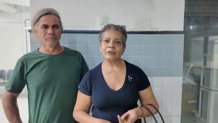 Mulher de cabelos curtos grisalhos e blusa azul-marinho aparece ao lado de um homem de cabelos grisalhos e camiseta verde, em ambiente interno com paredes revestidas de azulejos
