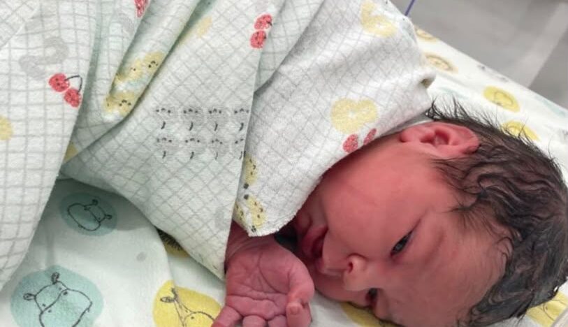 Bebê recém-nascida deitada de lado em superfície com estampas infantis, parcialmente coberta por manta clara, com o rosto e uma das mãos visíveis