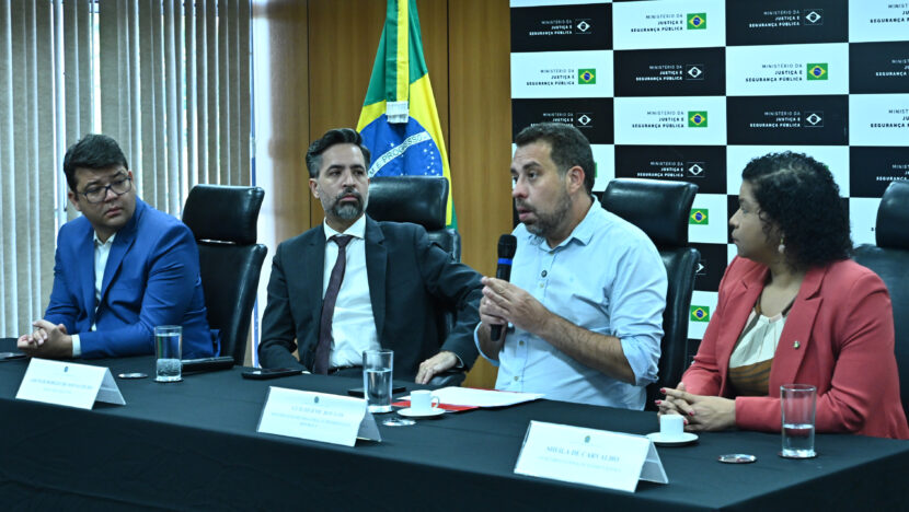 Integrantes do governo sentados à mesa em coletiva no Ministério da Justiça, com bandeira do Brasil ao fundo